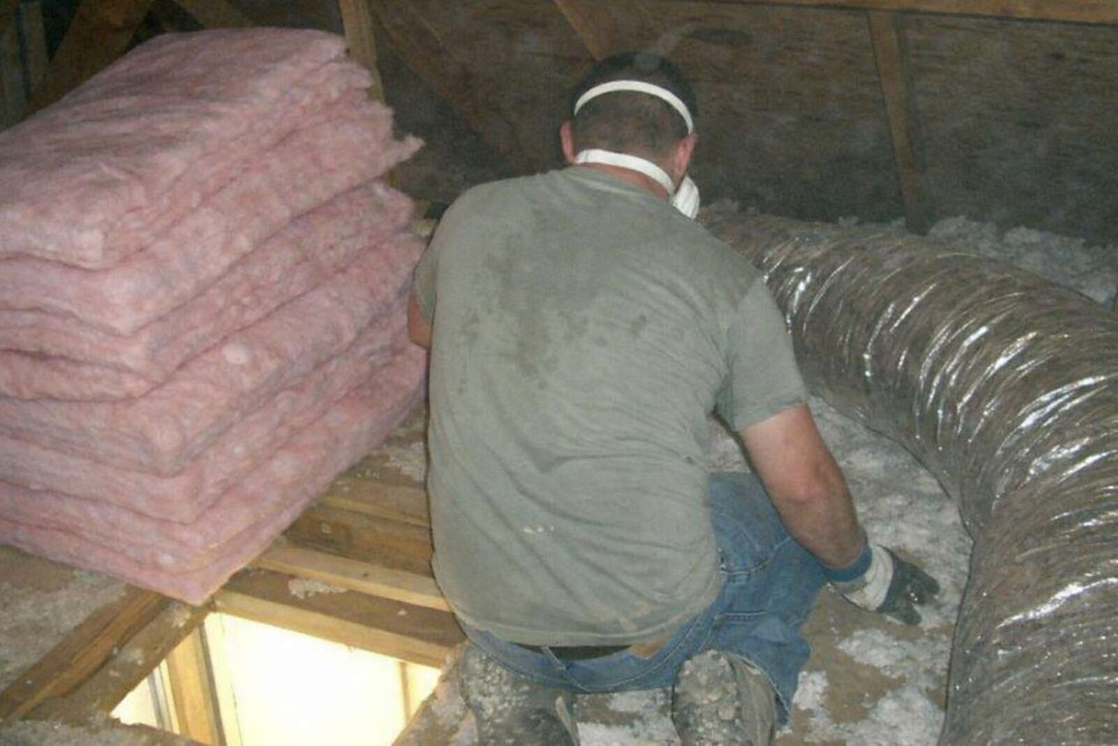 A gloved worker kneeling beside insulation batts in an attic, face out of frame